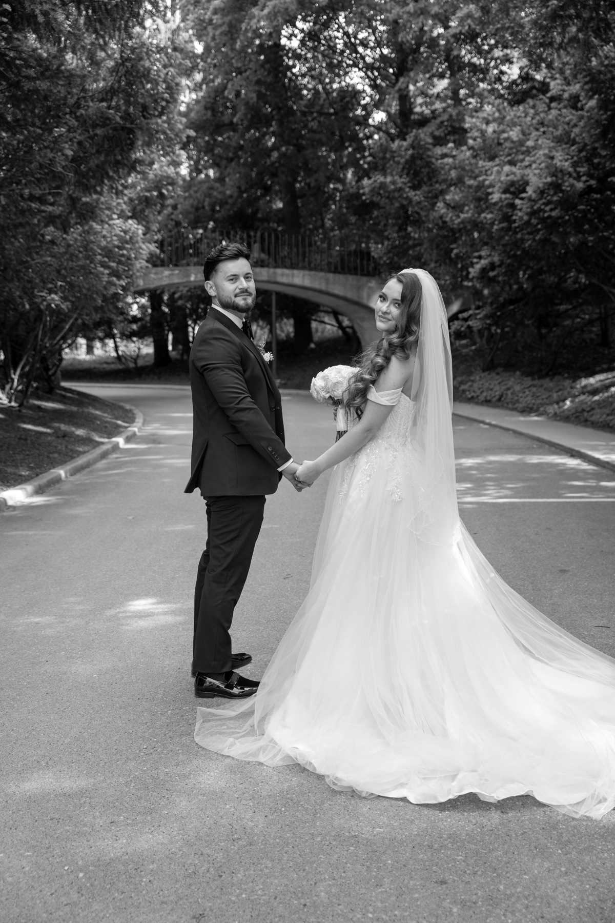 Black-and-white wedding portrait of a couple holding hands on a tree-lined path